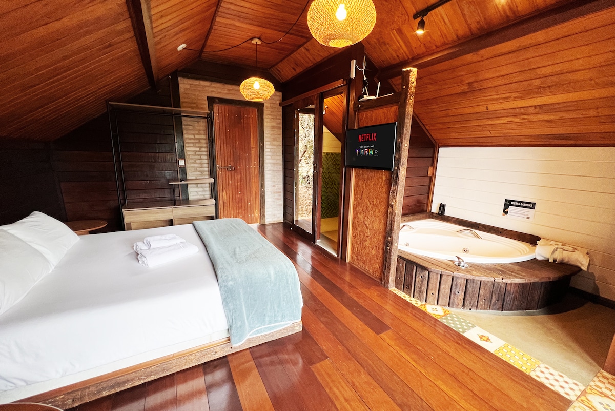 A spacious sleeping area features a Queen bed with a soft white duvet and a blue blanket draped across. A private hot tub, crafted from wood, is positioned nearby. Soft lighting from decorative fixtures enhances the natural wood accents of the room.
