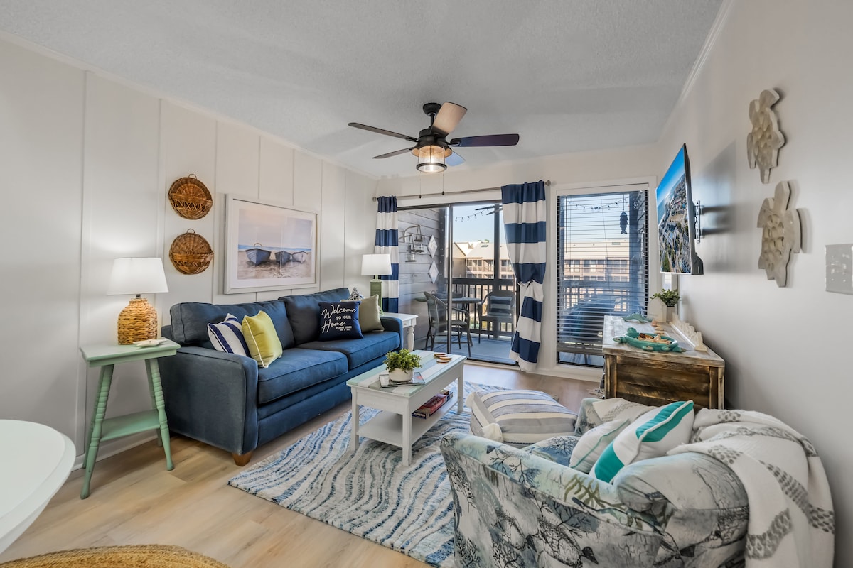 A bright living area features a comfortable blue sofa accented with decorative pillows. Two side tables hold lamps, and a unique art piece decorates the wall. Large glass doors open to a balcony, while natural light fills the space. A patterned area rug adds texture to the floor.