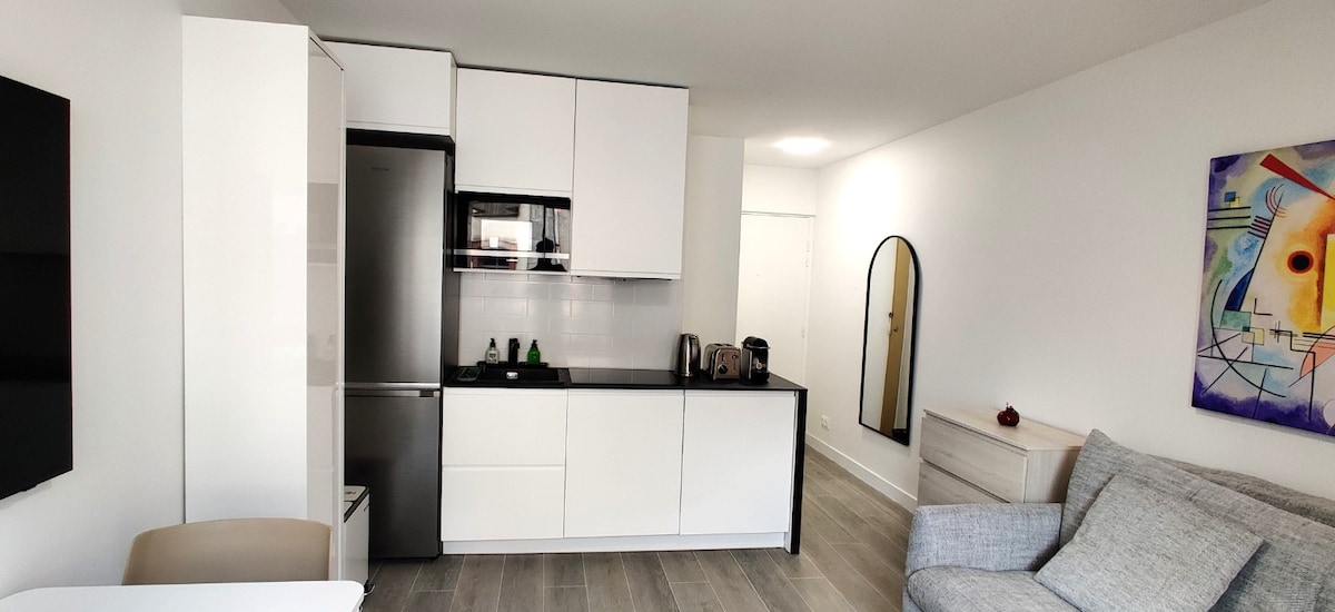 A modern kitchen area is equipped with sleek white cabinetry, a stainless steel refrigerator, and an induction cooktop. Light-colored flooring complements the bright walls. A mirror and a piece of colorful wall art add character, while a cozy gray sofa invites relaxation.