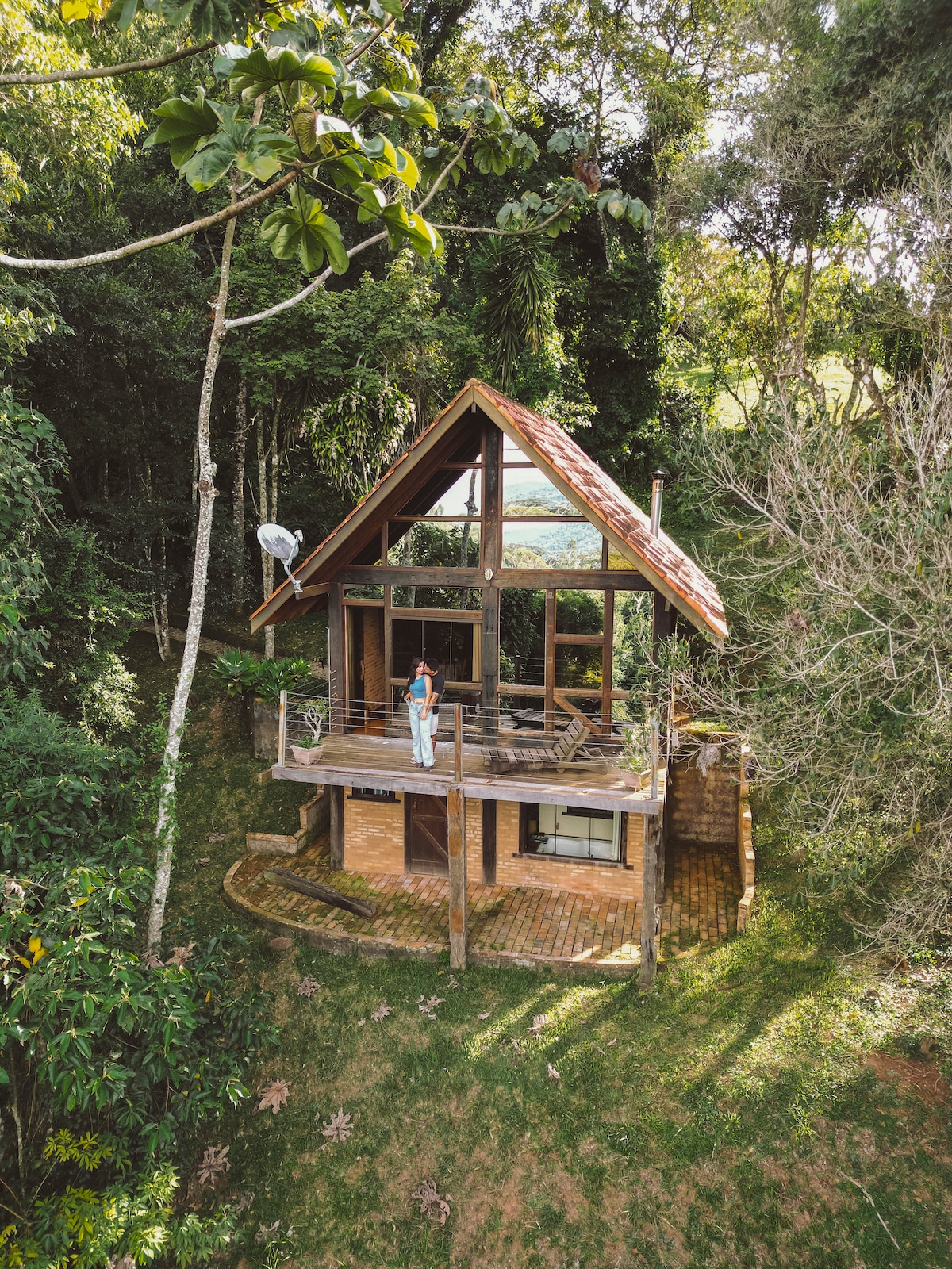 The cabin is set amidst lush greenery, featuring a sloped roof with wooden beams. A spacious deck is accessible, furnished with outdoor seating, and offers views of surrounding nature. Tall trees frame the structure, providing a sense of seclusion.