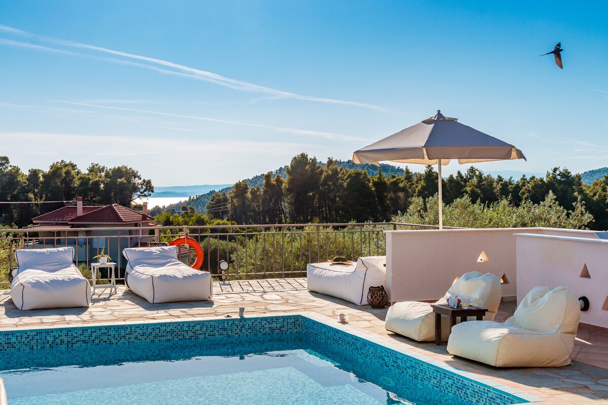 An outdoor area features a private swimming pool surrounded by sun loungers. A large umbrella provides shade over a seating area. The backdrop displays lush green pine trees and a view of the sea under a clear blue sky.