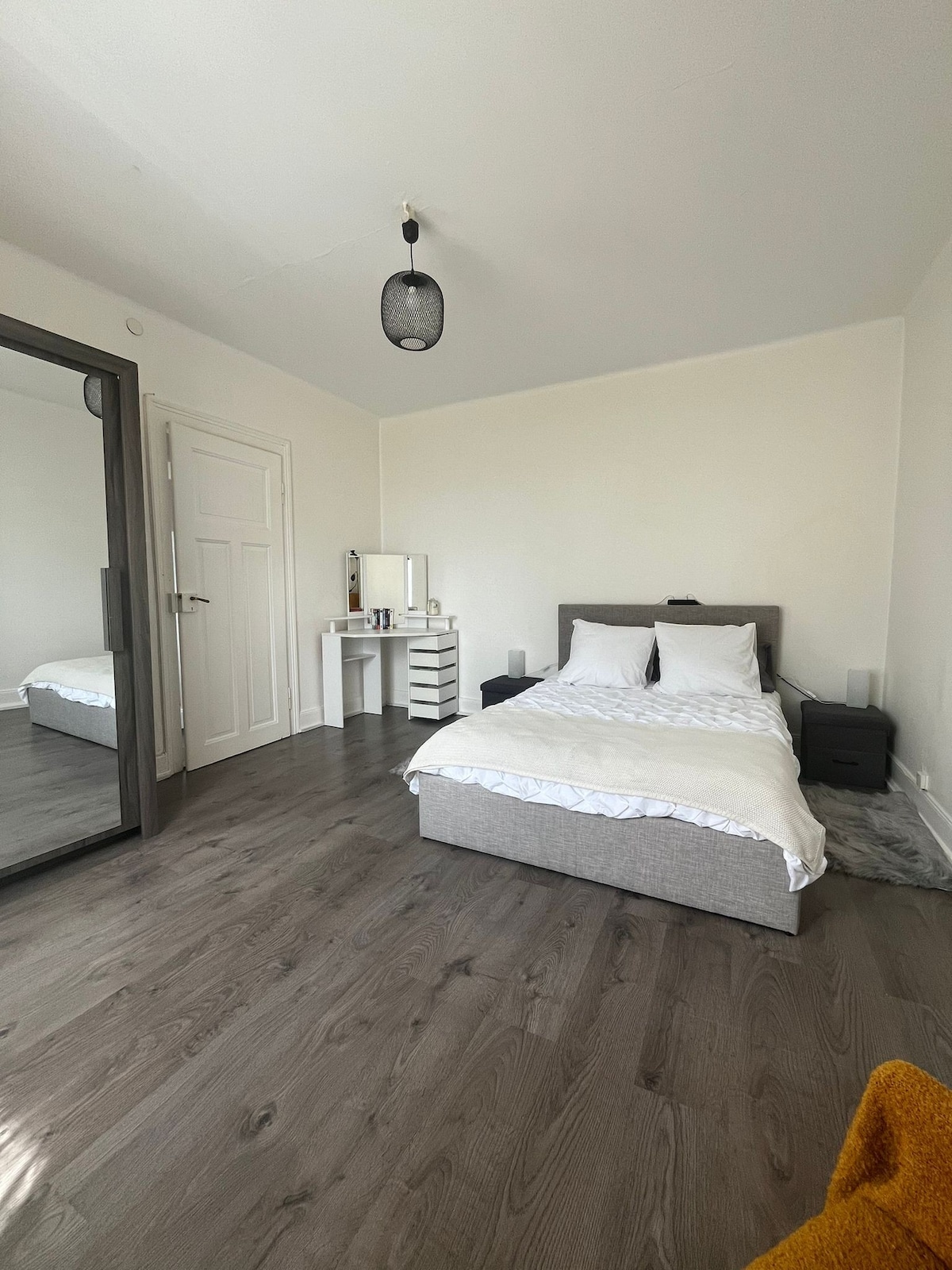 A simple bedroom features a spacious bed with white linens and two pillows. A large mirror reflects the room, enhancing the sense of space. A small desk with a chair and storage drawers is positioned nearby, complementing the neutral color palette and hardwood floor.