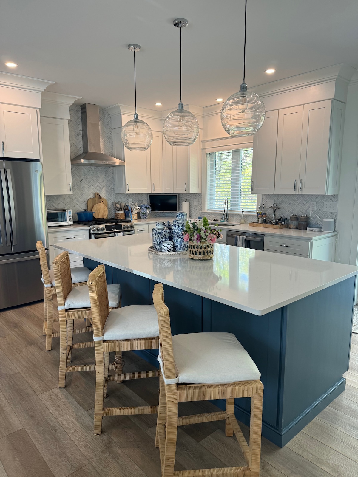 A modern kitchen showcases a large island with four counter-height chairs. Pendant lights hang above the island, illuminating the white and navy cabinetry. Stainless steel appliances and a marble backsplash complement the bright, airy atmosphere. A circular tray of fresh flowers adds a decorative touch.