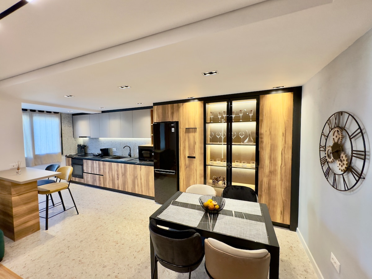 A modern kitchen and dining area is presented, featuring wood and dark cabinetry. A round dining table is set with four chairs, while a refrigerator and a glass-fronted cabinet provide ample storage. Natural light filters through the window, illuminating the spacious layout.