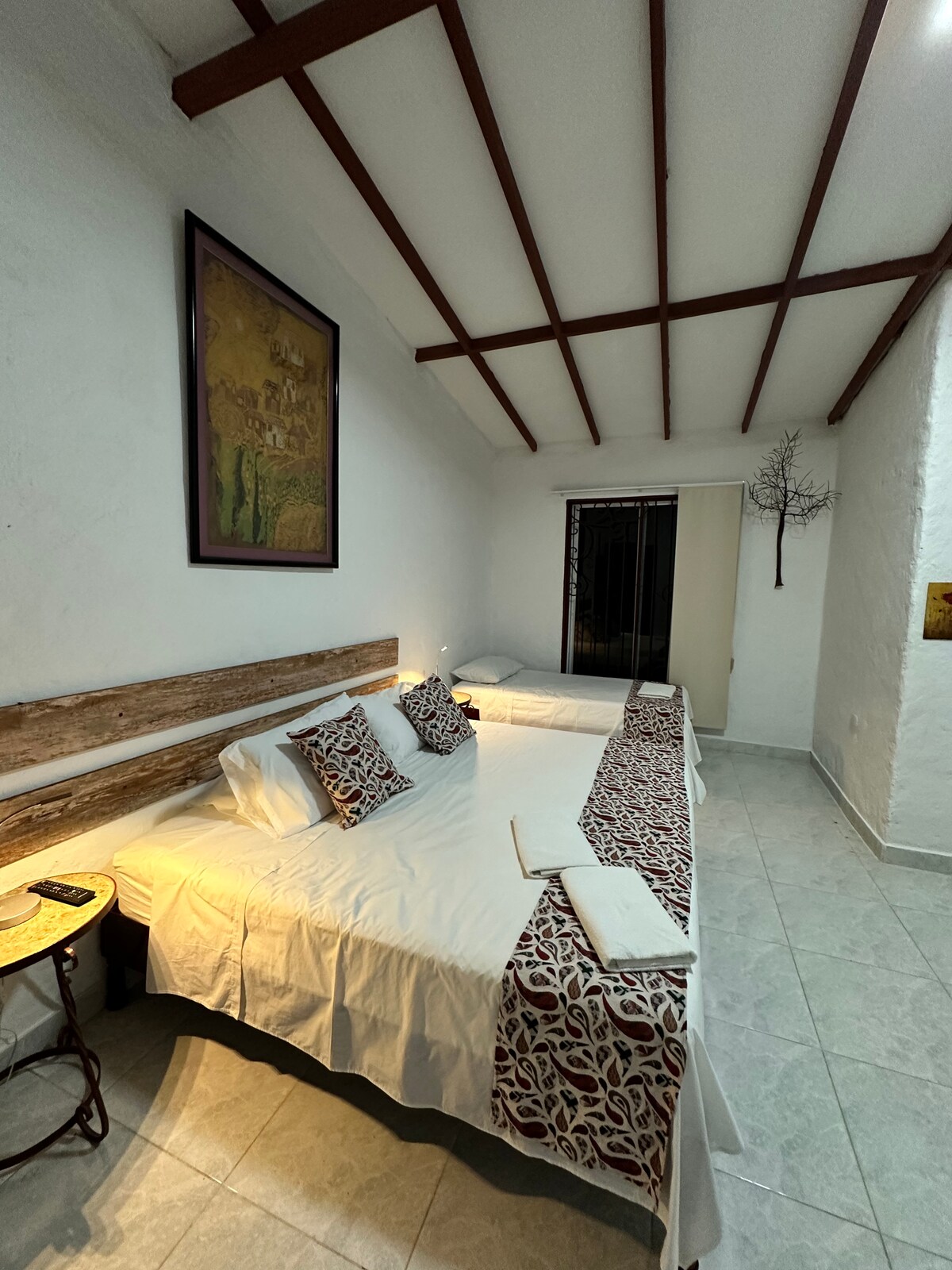 A cozy sleeping area is featured, containing a spacious double bed and two single beds. Decorative pillows add a touch of warmth. Soft lighting enhances the room's welcoming feel, which is complemented by a large window with curtains, allowing for natural light.