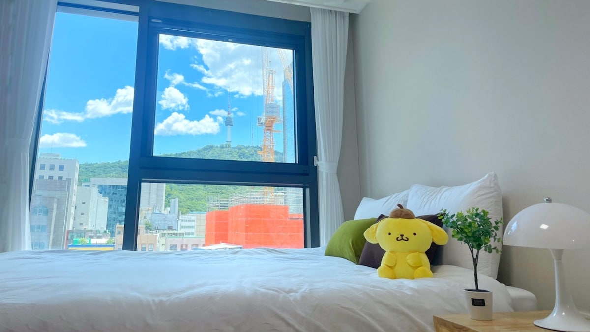A bright bedroom features a spacious bed adorned with soft pillows. A large window offers a view of the cityscape and mountains. A small plant and a lamp are placed atop a wooden nightstand beside the bed, adding a touch of comfort.