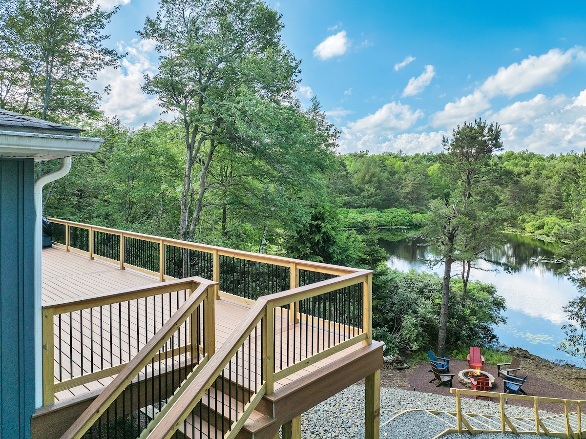 An expansive wooden deck offers a clear view of the surrounding greenery and a peaceful lake. A seating area with chairs and a fire pit is visible on the ground level, framed by lush trees under a clear blue sky.