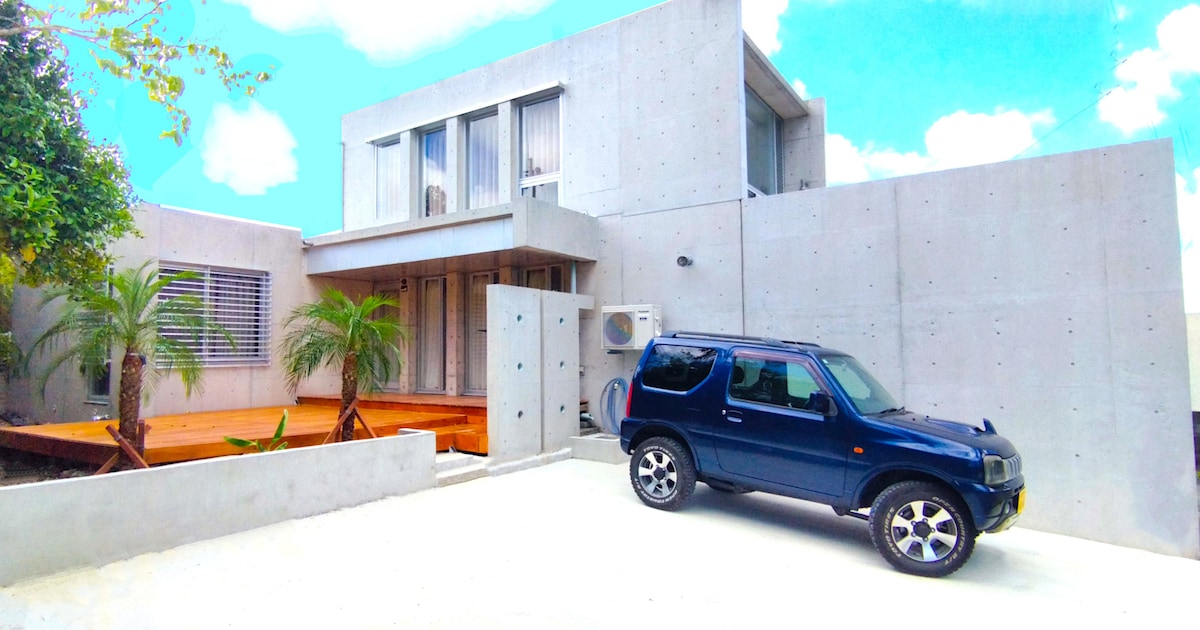 A concrete exterior features a modern design with large windows, allowing natural light to fill the space. Lush palm trees are present in the front yard alongside a parked blue SUV, creating a welcoming entrance to the property.
