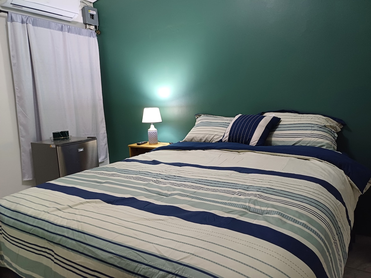 A cozy sleeping area features a spacious bed with a striped comforter, accented by decorative pillows. A small bedside lamp casts a gentle light, while a curtain partially covers a nearby window. A mini-fridge is visible in the corner, enhancing convenience.