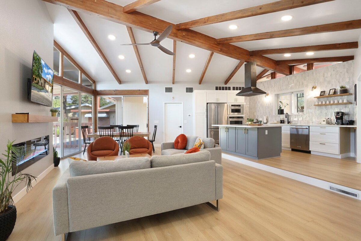 An open family room showcases high, exposed beam ceilings and an abundance of natural light. A modern kitchen features an island and sleek appliances, while a cozy seating area includes a light gray sofa and orange accents. Large windows provide views of the outdoor space.
