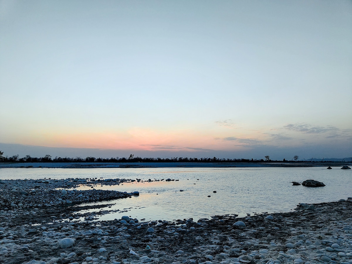 A serene waterway is captured at dusk, with gentle ripples reflecting soft hues of orange and pink in the sky. Smooth, rounded stones line the water's edge, contributing to the calm landscape that is surrounded by silhouettes of distant trees.