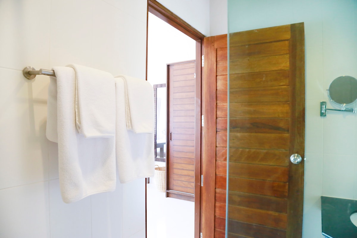 A well-lit bathroom showcases a wooden door leading to another space. Two neatly arranged towels hang on a stainless steel rack beside a glass-walled shower. The clean, white tiles create a fresh and inviting ambiance.