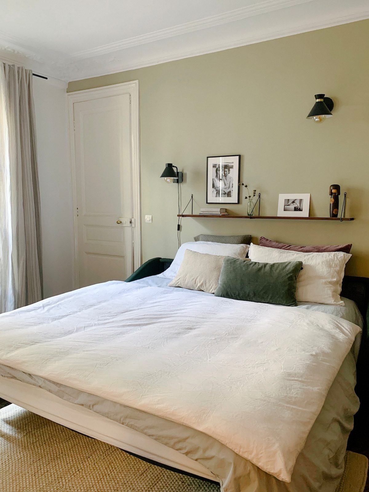 A cozy sleeping area is showcased with a spacious bed adorned with layered blankets and decorative pillows in soft tones. A small shelf displays framed photographs and decorative items. Natural light enters through a window dressed in light curtains, complementing the muted green wall.