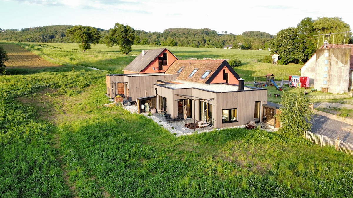 An architectural design featuring a spacious house set in a natural landscape. The exterior showcases multiple sections with a mix of wood and maroon facades. Surrounding green fields and trees contribute to its serene setting, providing an inviting atmosphere for gatherings.