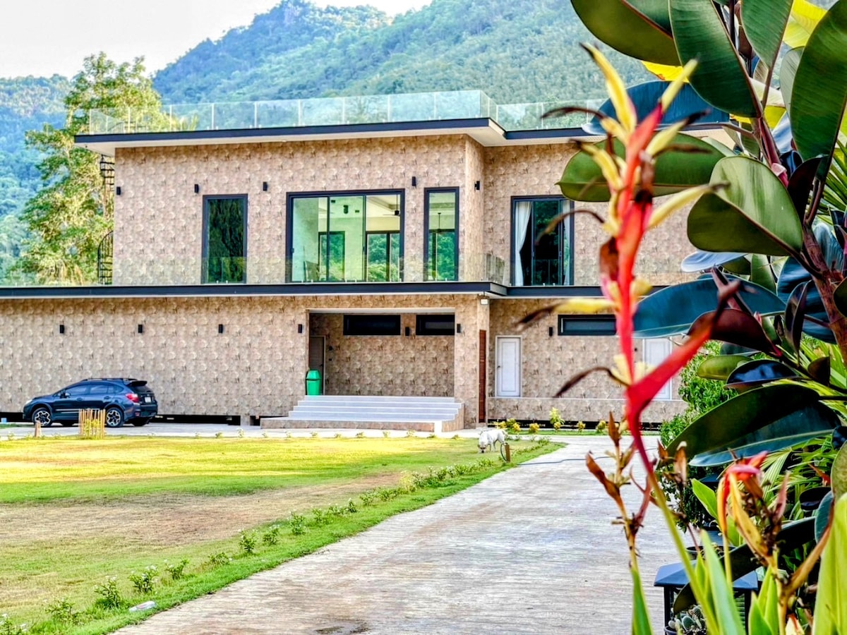 A modern two-story building is displayed against a backdrop of lush mountains. The façade features stone-patterned walls and large glass windows. A driveway leads to the entrance, accompanied by a neatly landscaped yard with colorful plants framing the scene.