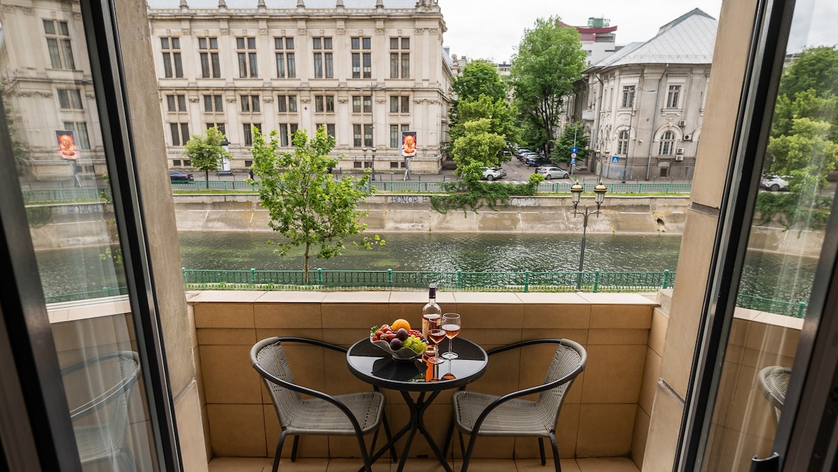 A balcony offers a cozy seating arrangement with two chairs and a round table. A bottle of wine and a bowl of fresh fruit are elegantly displayed, while an unobstructed view of the Dâmbovița River and surrounding greenery can be seen in the background.