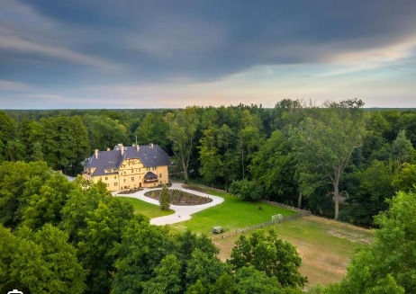 A stately mansion is surrounded by lush greenery, showcasing a well-manicured lawn and a circular driveway. The structure's distinct architecture is highlighted against the backdrop of a vast forest, creating a serene atmosphere ideal for relaxation.