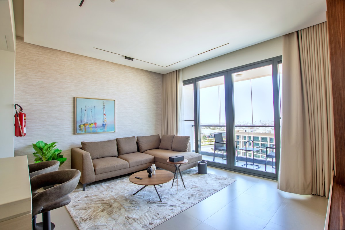 A spacious living area is revealed, featuring a comfortable sofa and a circular coffee table. Large windows allow natural light to fill the room, with a view of the outdoors visible. A piece of art adorns the wall, and a small plant adds a touch of greenery.