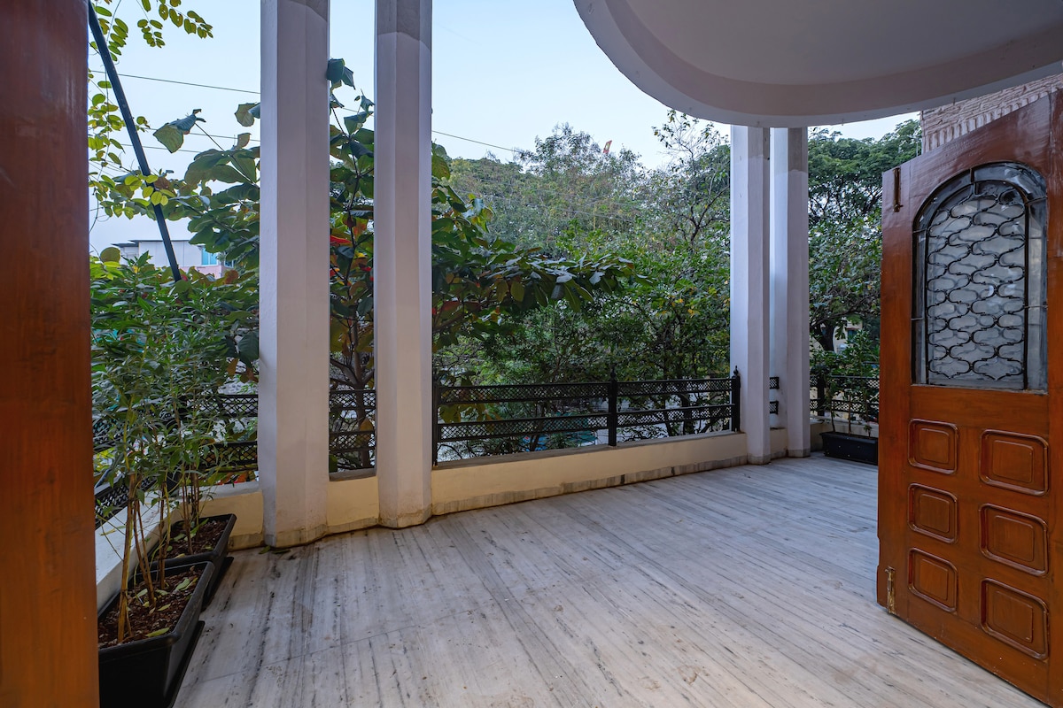 A spacious balcony is framed by tall columns, offering views of lush greenery. Potted plants add a touch of nature, while the wooden door to the interior provides a warm entrance. The floor is finished in light wood, enhancing the open and airy feel.