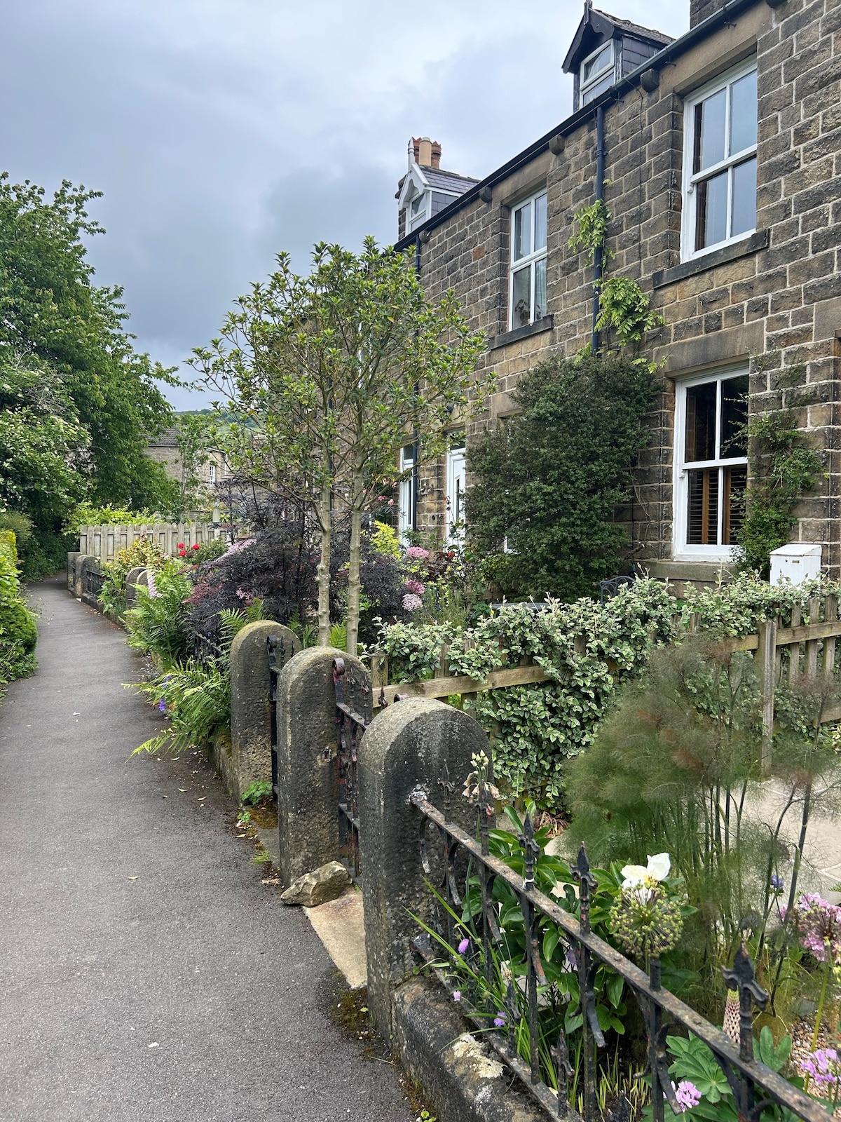 A charming terrace house is framed by a well-maintained garden, featuring a variety of plants and flowers. A gravel path runs alongside the property, bordered by a low stone wall. Trees and shrubs provide lush greenery, with windows partially obscured by foliage.