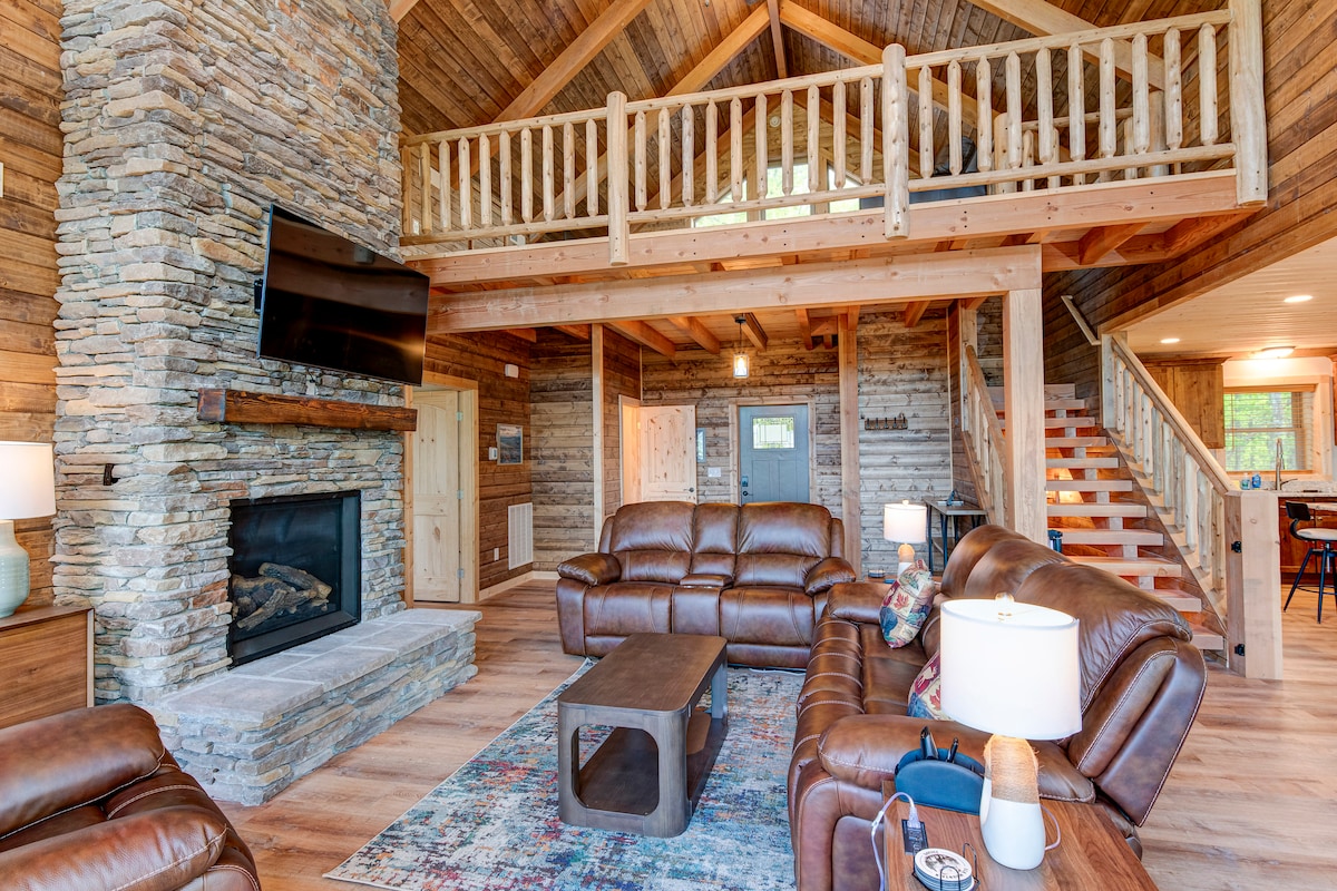 A spacious living area is adorned with natural wood accents and a stone fireplace. Comfortable leather sofas are positioned around a wooden coffee table. A loft area is visible above, with a wooden railing overseeing the open space. Large windows let in natural light, enhancing the warm ambiance.