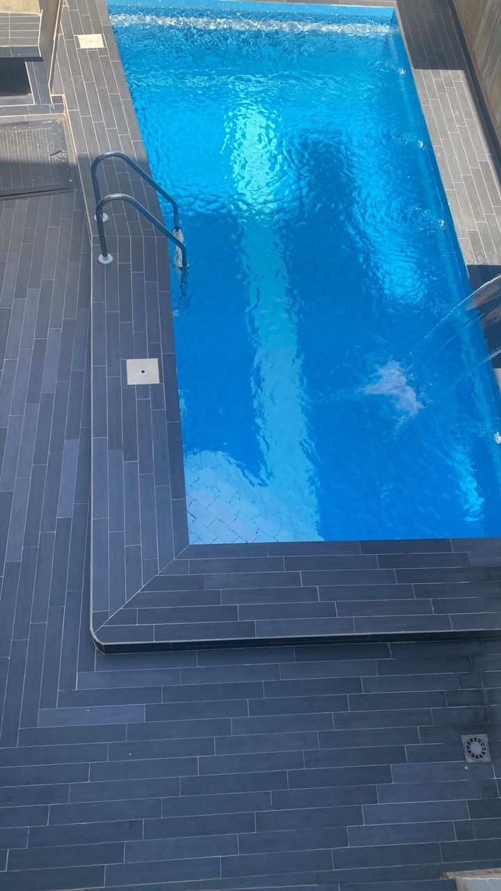 The image showcases a serene swimming pool with clear blue water, surrounded by sleek dark tile flooring. A single ladder leads into the pool, and the sunlight creates soft reflections on the water's surface, enhancing the tranquil setting.