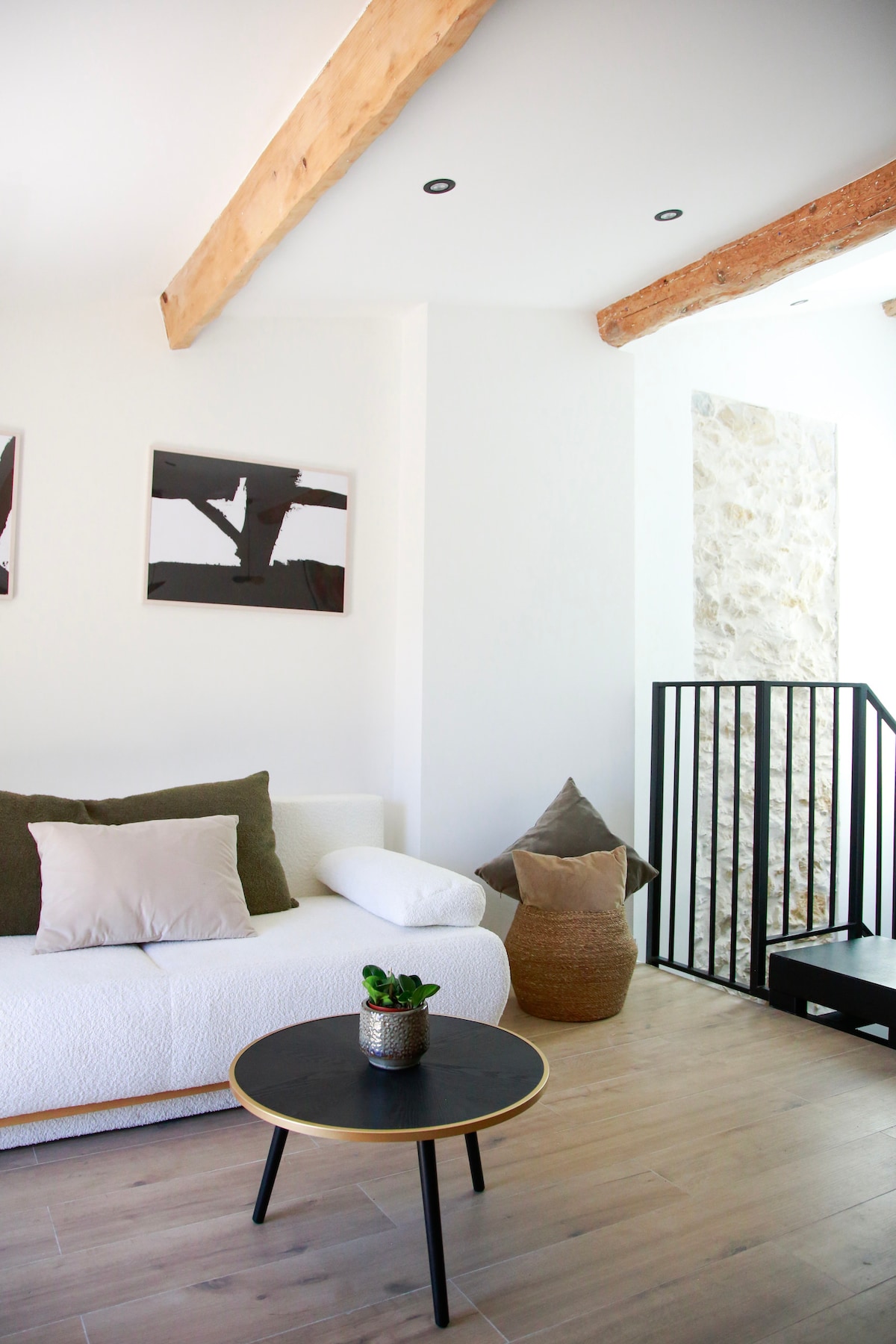 A bright living area features a cozy white couch adorned with soft cushions. Two framed artworks are displayed on the walls, and a decorative potted plant rests on a simple black table. Exposed wooden beams and a stone wall add to the inviting ambiance.