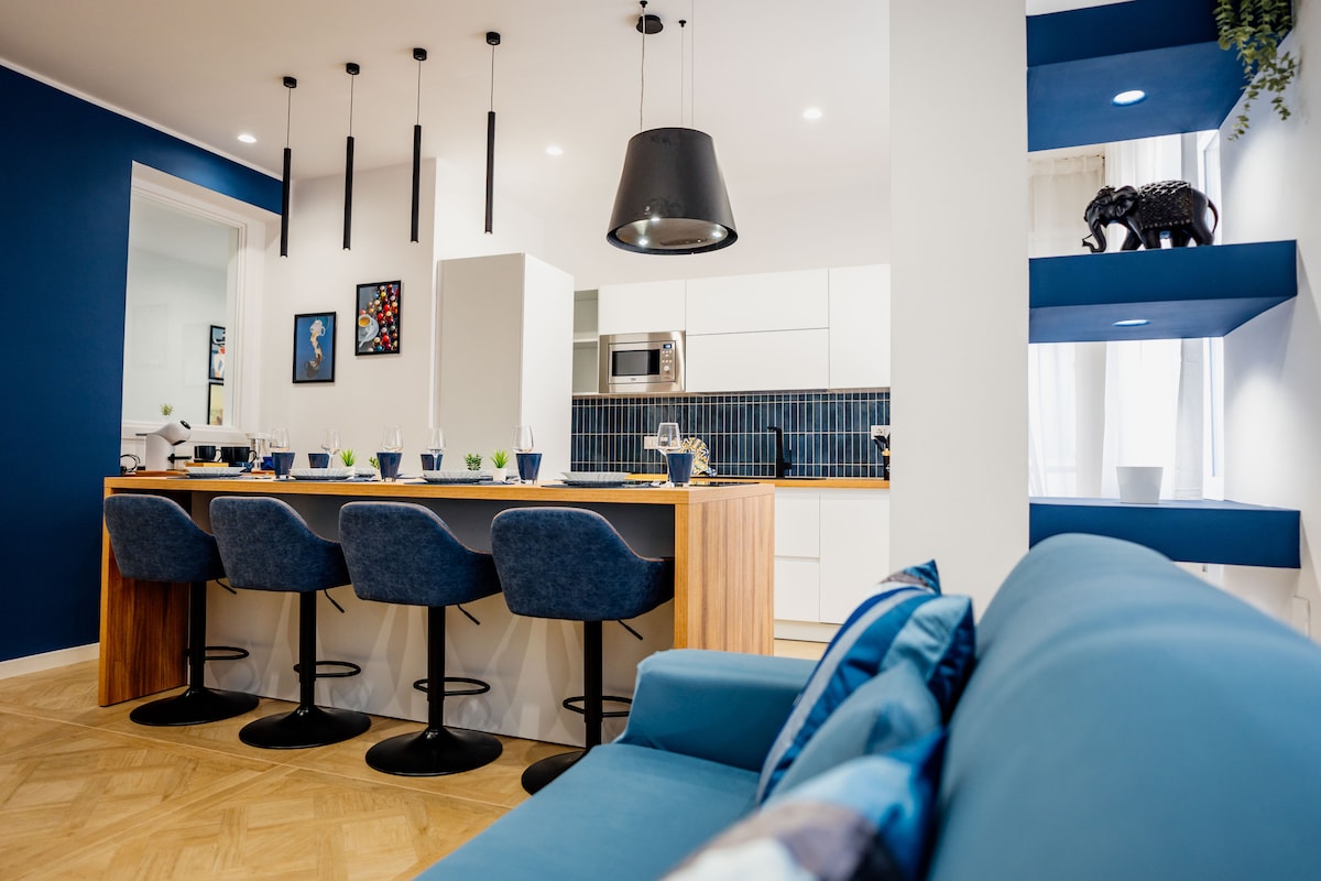 A modern kitchen space is showcased with a central island featuring seating for four. Stylish blue and white decor is highlighted by pendant lighting. A comfortable sofa in the foreground invites relaxation, while decorative elements add a touch of personality.