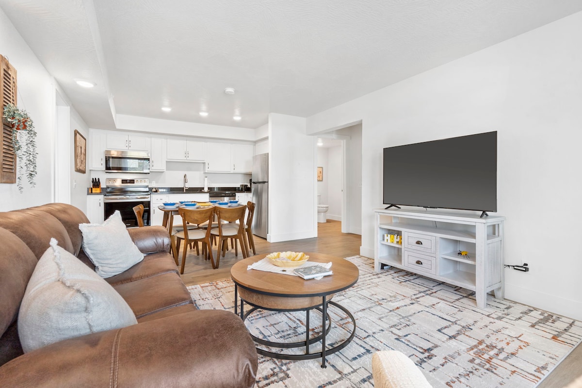 A spacious living area features a cozy brown couch accompanied by decorative pillows. A coffee table sits in the center, surrounded by a stylish area rug. In the background, a dining table for six is visible next to a well-equipped kitchen, with a television mounted on a light-colored media console.