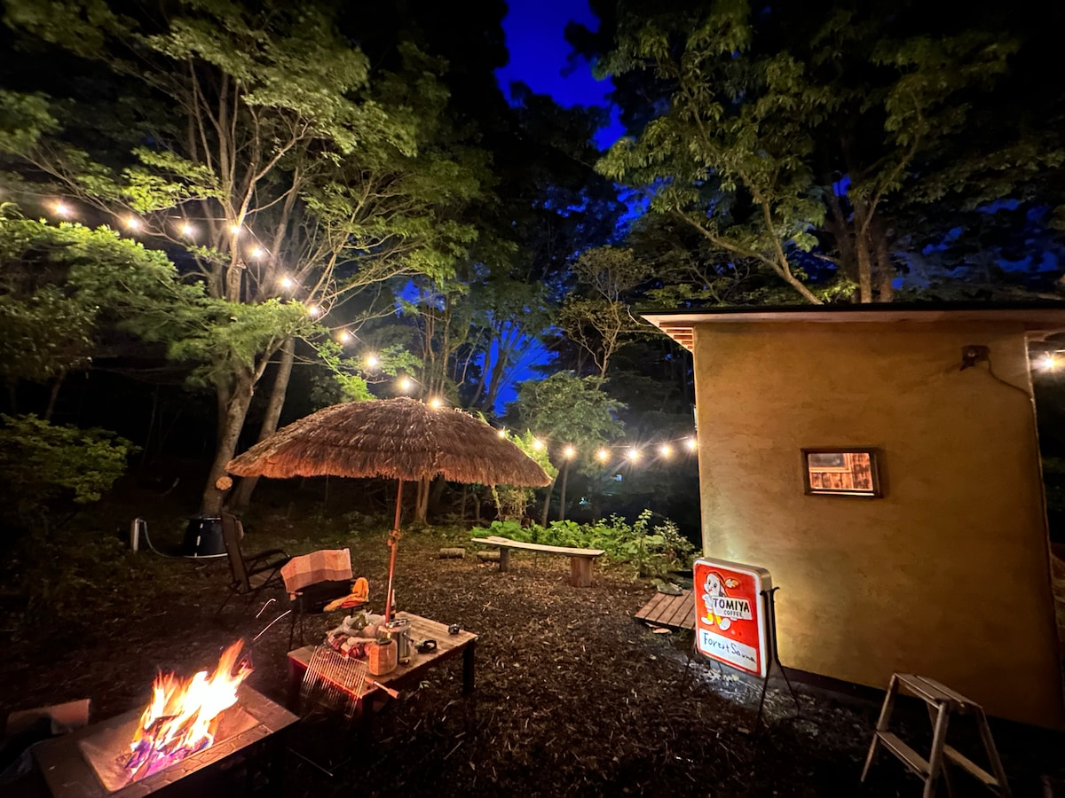 An outdoor area is illuminated by soft string lights hanging above, creating a cozy ambiance. A thatched umbrella provides shade over a seating area, while a small fire burns on the table. Lush greenery surrounds the scene, enhancing the tranquil atmosphere.