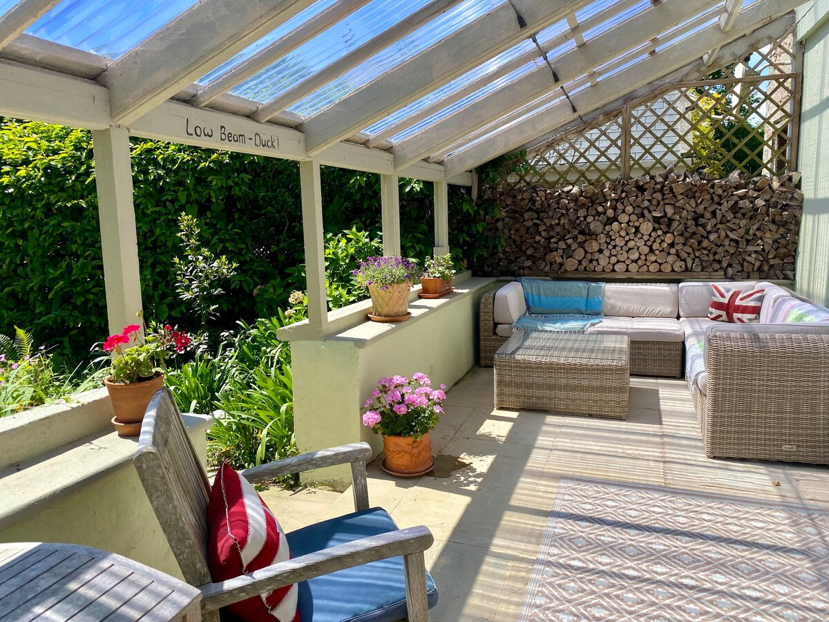 A covered terrace is presented, featuring a comfortable seating area with a sectional sofa and vibrant throw pillows. Potted plants add color to the space, while a rustic wood stack serves as a decorative backdrop. Natural light filters through the transparent roof.