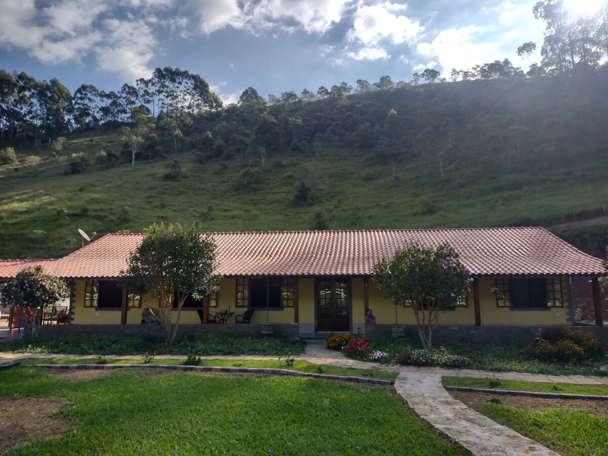 The property features a charming, single-story building with a terracotta roof, set against a lush green hillside. Surrounding the structure are well-maintained gardens with various plants, and a stone pathway leads to the entrance.