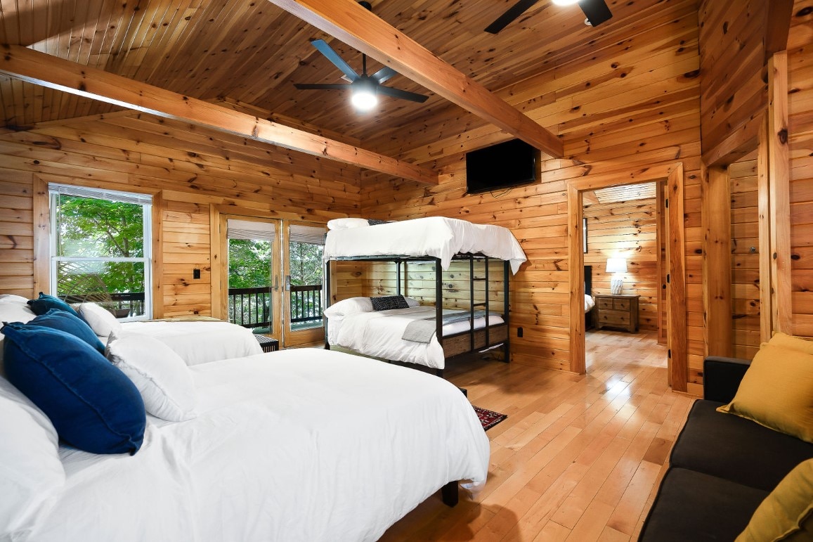The interior features a wooden design with vaulted ceilings and large windows. Two beds are situated in the space, including a bunk bed. Soft linens cover the beds, and a seating area is present with cozy cushions. Ceiling fans provide additional comfort.