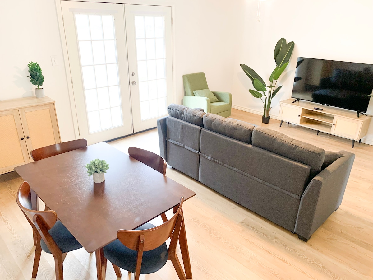 A spacious living area features a dark gray sofa facing a wall-mounted television. A light wooden dining table with four chairs is positioned nearby, complemented by a small potted plant at the center. A green accent chair and a tall indoor plant add a touch of color.