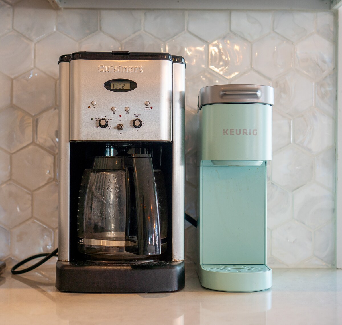 A modern coffee preparation area features a Cuisinart coffee maker alongside a stylish mint green Keurig machine. Both appliances are positioned against a textured backsplash of hexagonal tiles, enhancing the kitchen's functional design.