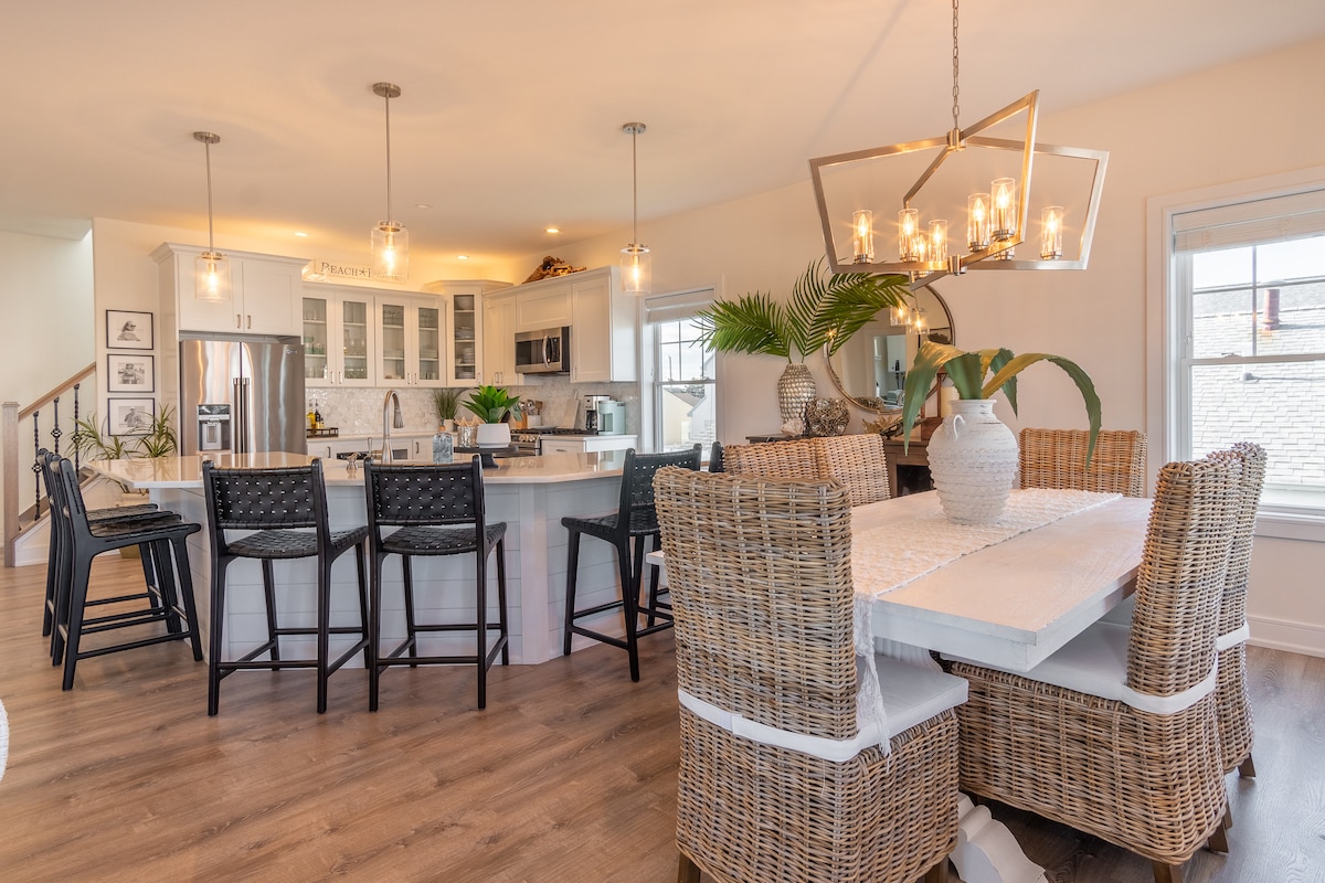 An open-concept kitchen features white cabinetry and stainless steel appliances, complemented by a large island with seating for six. A stylish dining table made of woven material accommodates six, while elegant light fixtures hang above, enhancing the modern, bright space filled with greenery and natural light.