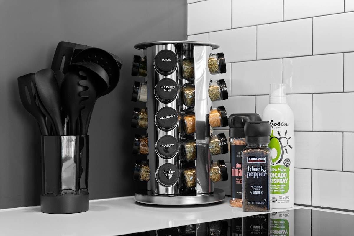 A rotating spice rack is prominently displayed on the kitchen countertop, alongside a container for cooking utensils. Various spice jars are organized around the rack, labeled with herbs and seasonings. Additional spice containers and a cooking spray are visible nearby, enhancing the kitchen's functionality.
