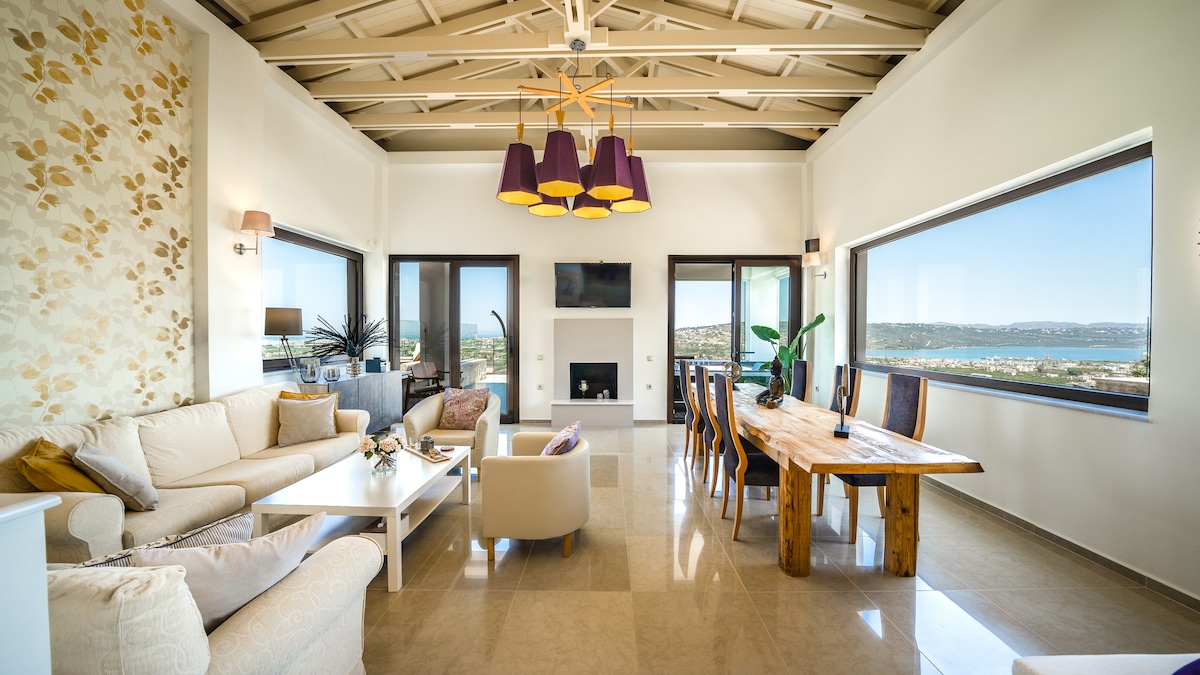 A spacious living and dining area features large windows that allow natural light to fill the room and showcase panoramic views of the ocean. Comfortable seating includes a light-colored sofa and armchairs, complemented by a wooden dining table surrounded by chairs. Modern lighting fixtures add a touch of elegance.