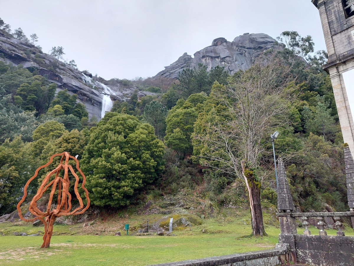 A scenic view features a large rocky cliff with a waterfall cascading down among dense greenery. A unique metal sculpture resembling a tree stands in the foreground. The surrounding landscape is lush, with trees providing a natural backdrop and a serene atmosphere.
