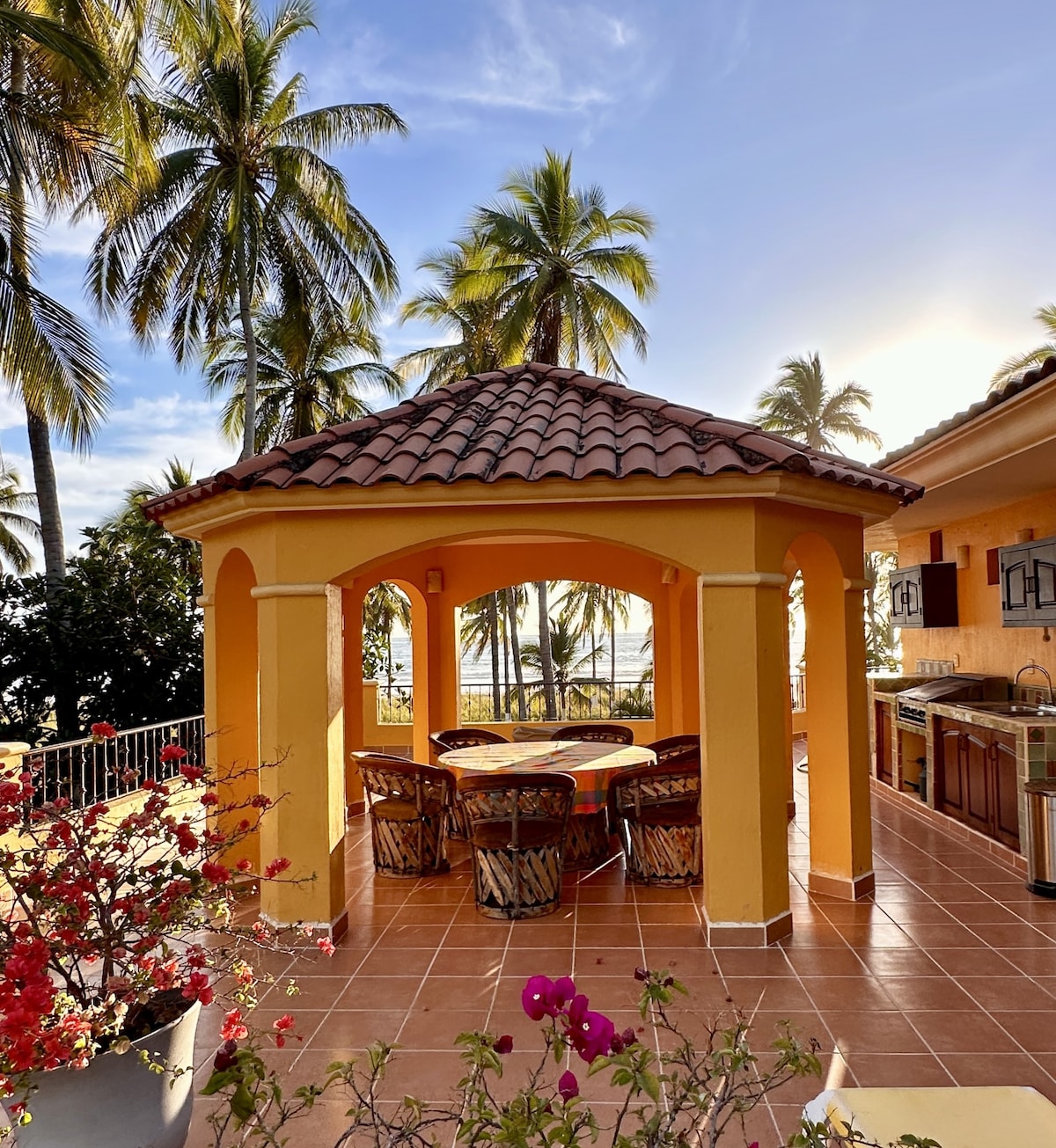 An outdoor palapa features a circular dining table surrounded by wicker chairs. The vibrant orange structure is framed by lush palm trees, with beach and ocean views visible in the background. Bougainvillea plants add a touch of color to the scene.