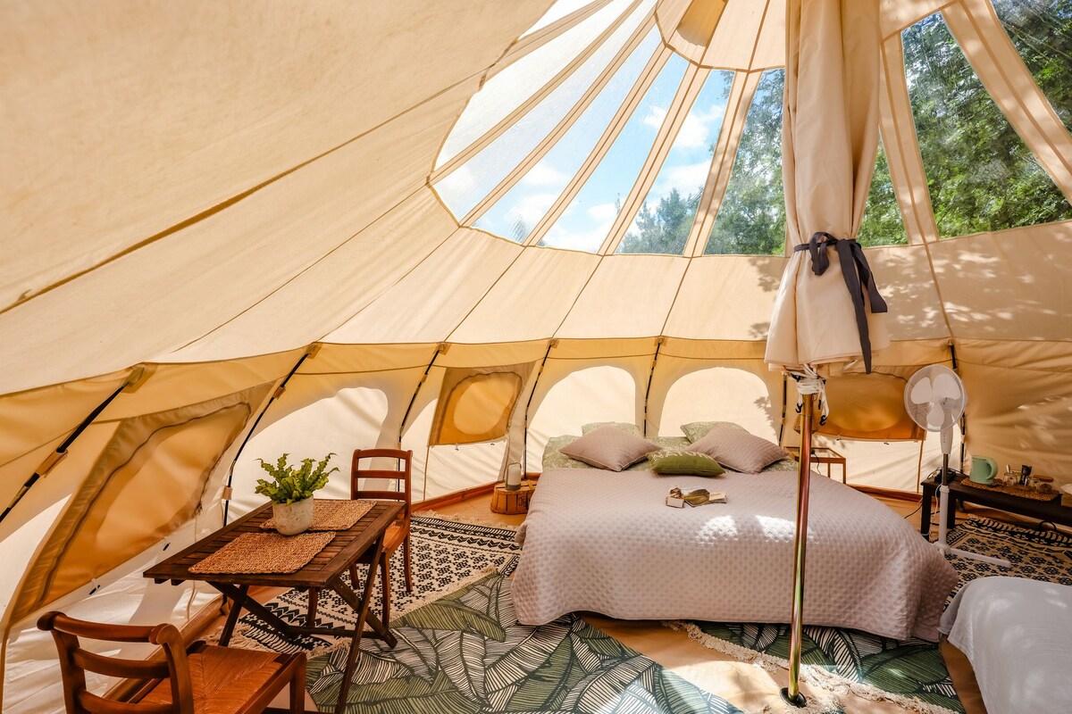 The interior of a Lotus Belle tent is shown, featuring a spacious bed adorned with neutral bedding. A wooden table and chairs are arranged nearby, complemented by a patterned rug. Natural light floods the space through the tent's large windows, highlighting the warm tones of the fabric.