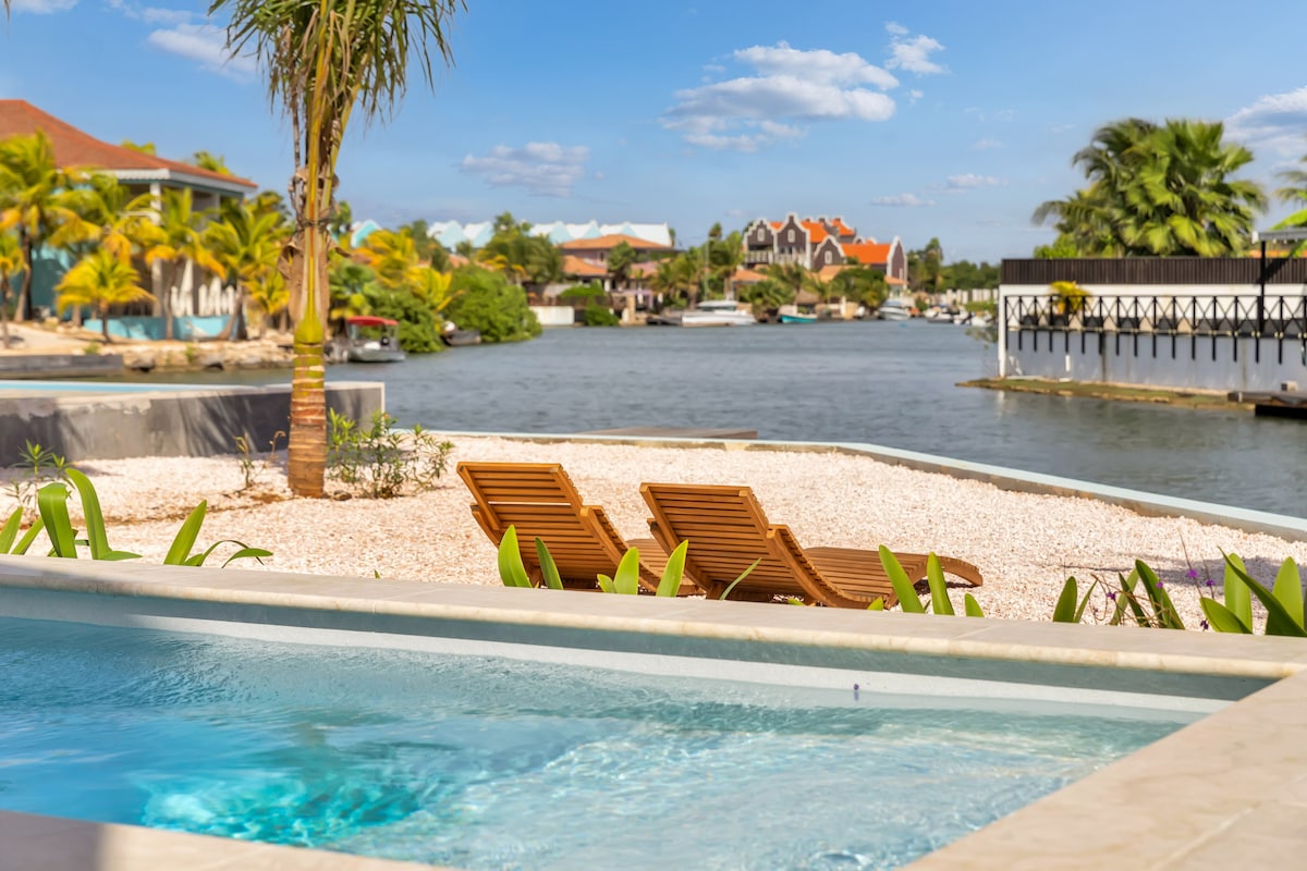 A private pool is positioned at the forefront, surrounded by a landscaped area with shrubs and palm trees. Two lounge chairs are placed nearby, offering a view of the calm lagoon and waterfront homes, with blue skies and scattered clouds in the background.