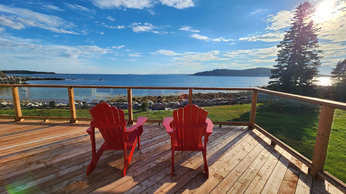 Two red Adirondack chairs are positioned on a wooden deck overlooking St Margaret's Bay. The view features a scenic coastline with rocky shores and trees in the background, illuminated by natural light under a blue sky with scattered clouds.