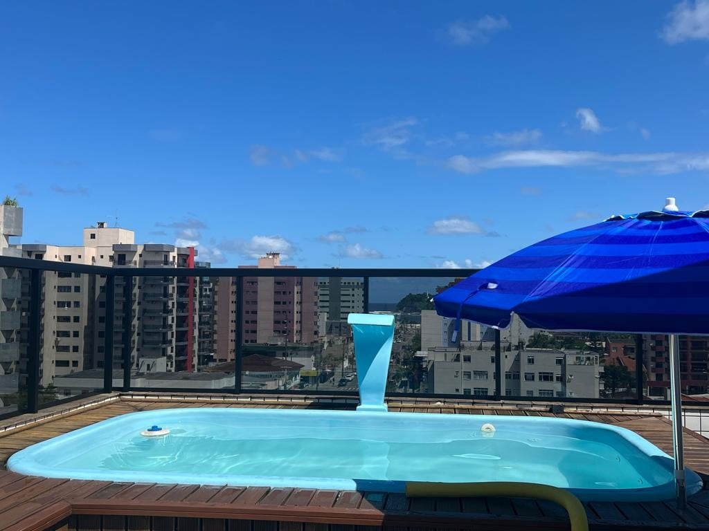 A private heated pool is seen atop a rooftop deck, surrounded by a glass-walled terrace. The clear blue water reflects the sky, while a large umbrella provides shade. A backdrop of city buildings can be seen, creating a serene urban oasis.