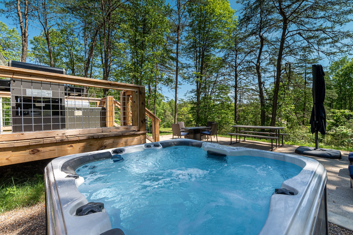 A relaxing hot tub is set under a clear blue sky, surrounded by lush greenery. Nearby, a gas grill is positioned on a wooden deck, accompanied by seated areas for outdoor dining and enjoyment.