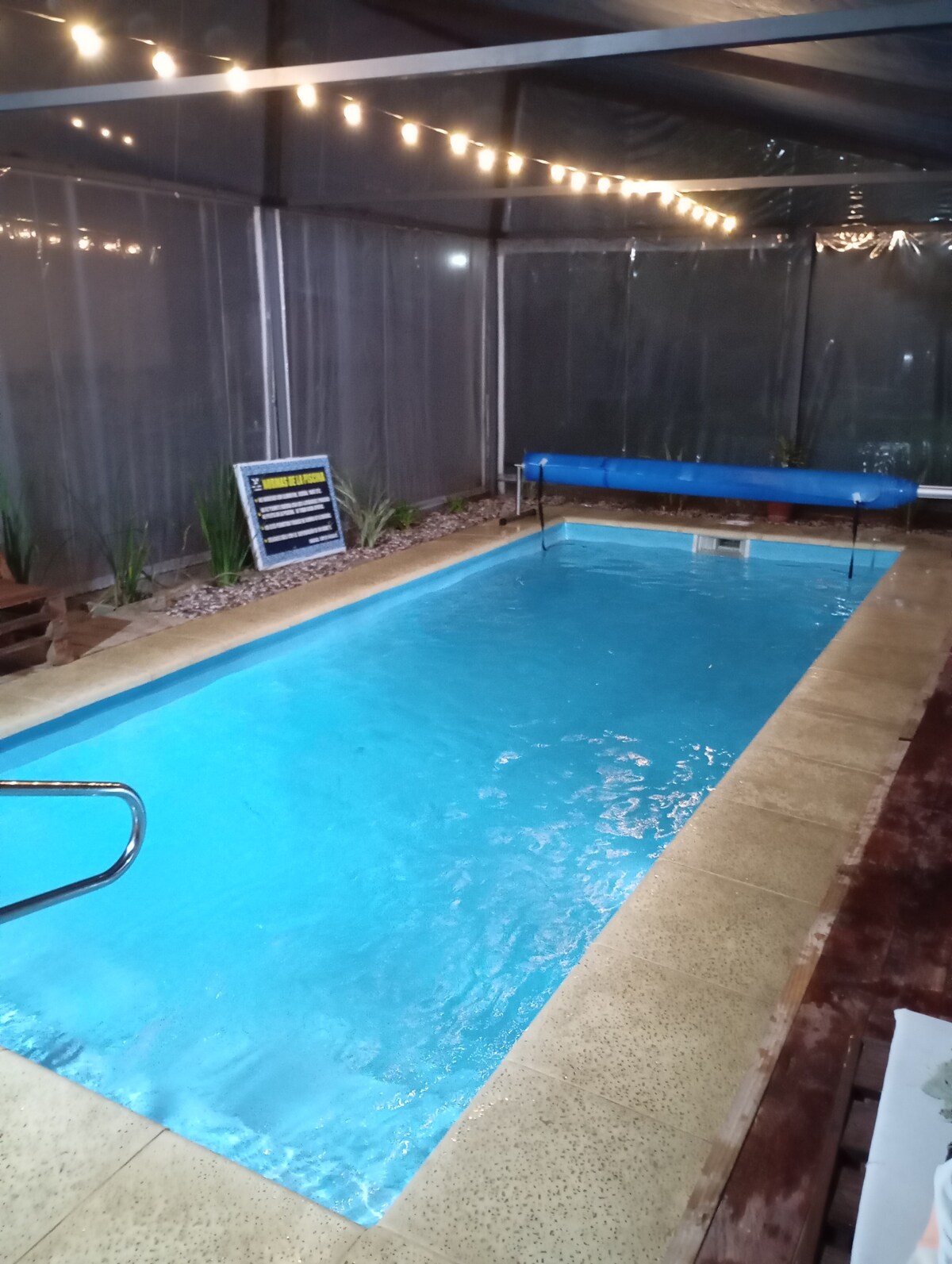 A privately enclosed heated pool is present within the facility, featuring a smooth concrete deck. Soft lighting is provided by string lights overhead, creating a serene ambiance. Small plant accents surround the pool area, enhancing the tranquil environment.