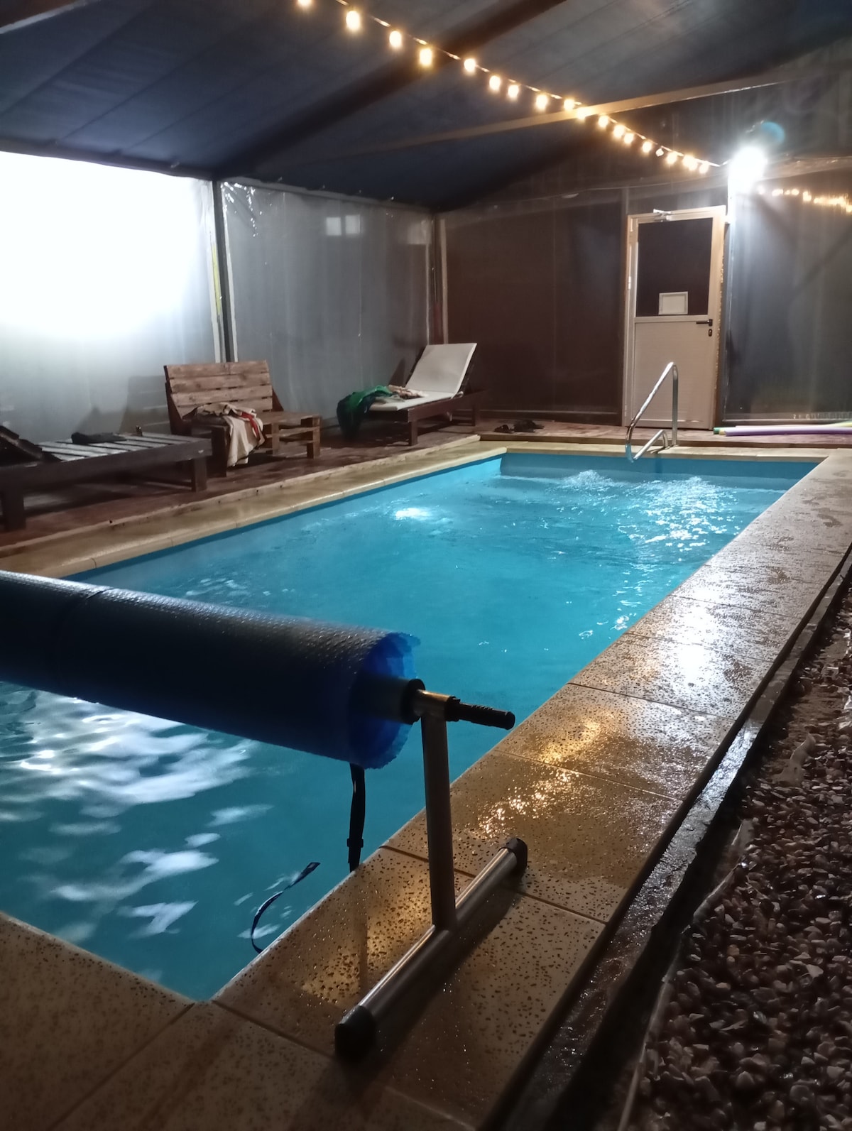 An indoor pool is surrounded by a tiled area, with a blue cover partially visible at the edge. Wooden lounge chairs are placed nearby, and subtle overhead lighting creates a calming ambiance. The entrance door is located in the background, framed by translucent privacy panels.