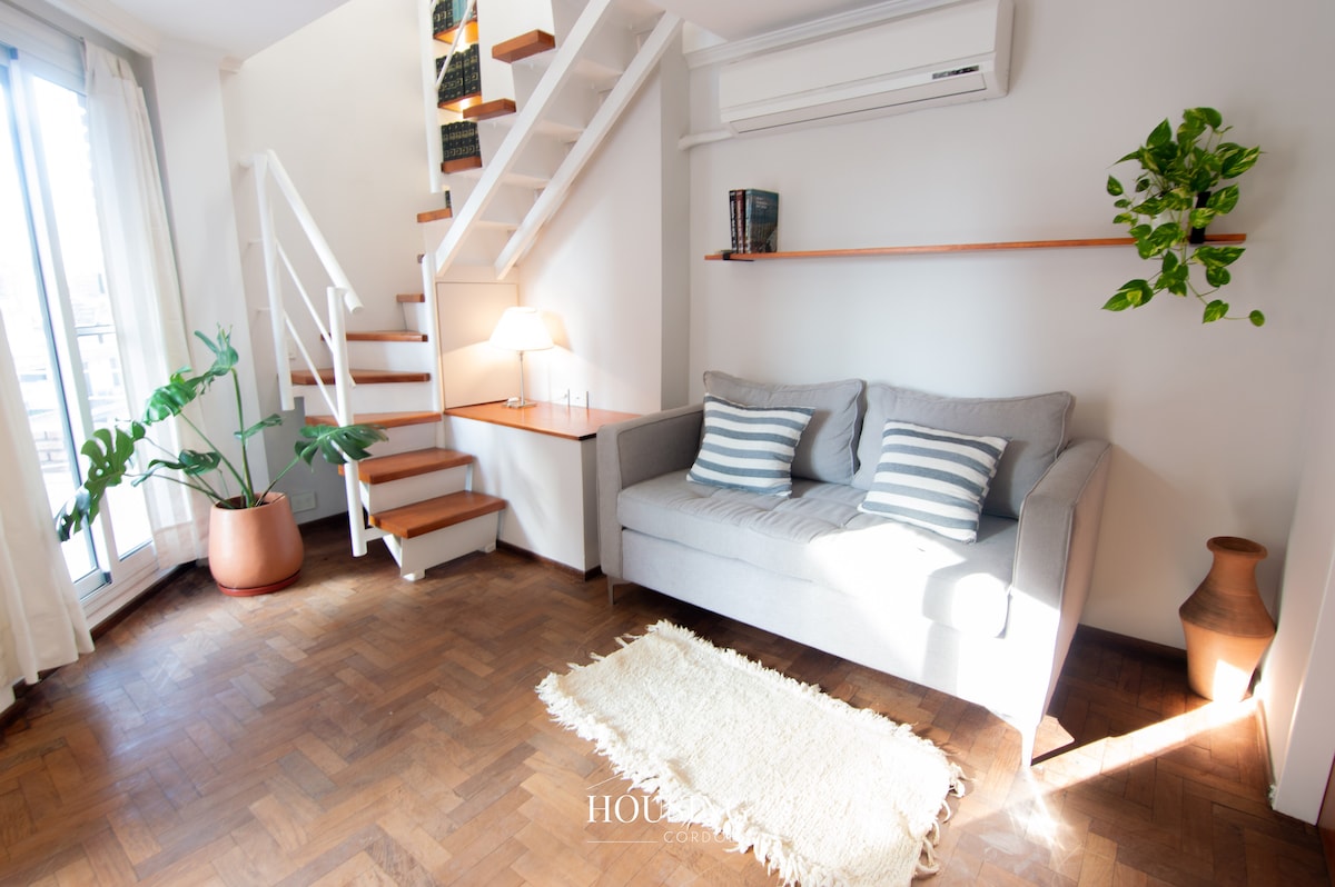 A modern living area features a light grey couch adorned with striped cushions. A soft woven rug lies on polished wooden floors. Natural light filters in through glass doors, illuminating the space. A small staircase leads to an upper area, complemented by indoor plants and a warm lamp.