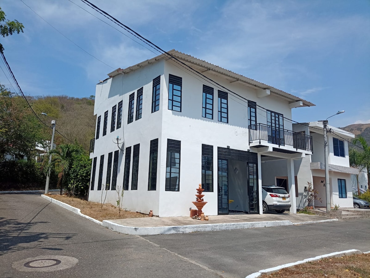 A two-story building with a light-colored exterior features numerous windows framed in black. The entrance is marked by a simple pathway leading to a small decorative fountain. A vehicle is parked in the driveway, under a clear blue sky.