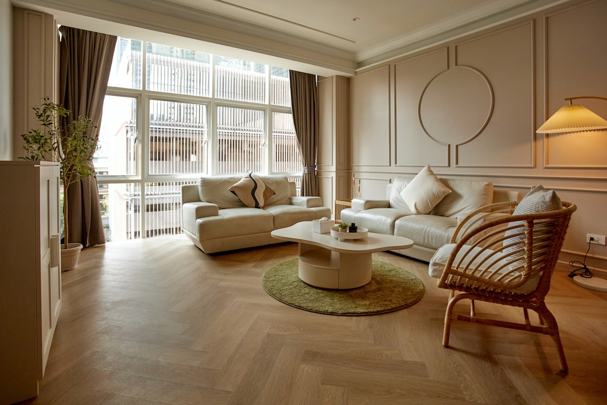 A cozy living area is highlighted by two light-colored sofas and a round coffee table, surrounded by large windows with soft curtains. A natural fiber rug lies on the floor, while a decorative lamp and potted plants add elements of warmth.